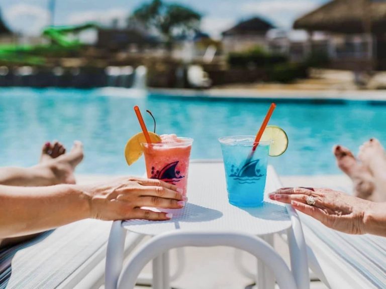 Pink and blue margaritas on a table with two hands reaching towards them by a resort pool