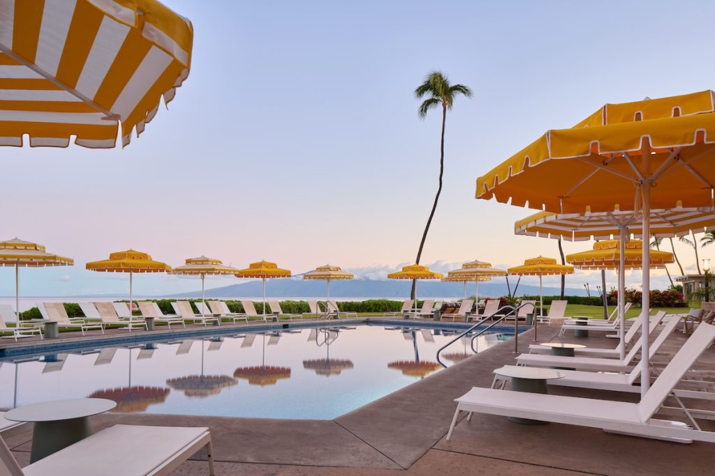 Yellow and white striped umbrellas with white chaise lounges surround the pool at the Royal Lahaina Resort & Bungalows on Maui.